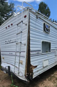 A white RV with a broken window and a ladder leaning against it, parked in an outdoor setting.